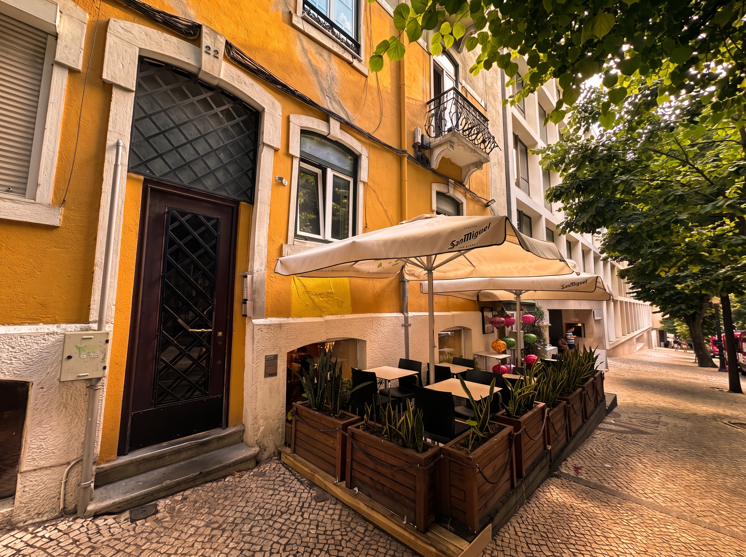 Street facade of Rua Camilo Castelo Branco 22 in Lisbon at sunset, with café terrace under the trees